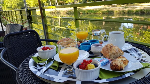 A breakfast setup on a table with a view of a lake, including a glass of orange juice, a jug of milk, pastries, and fruit. - Péniche Amboise Maison d'hôtes Toulouse (Toulouse)
