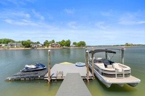 A dock with a pontoon boat and a jet ski, surrounded by calm water and houses in the background.