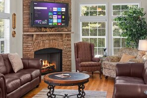 A living room with a fireplace, leather sofas, a round coffee table, and a flat-screen TV mounted above the fireplace displaying a streaming service interface.