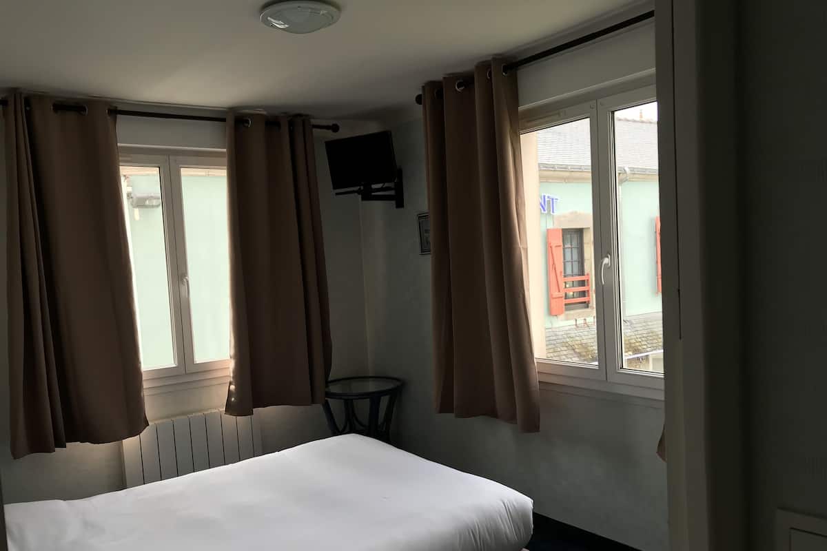A hotel room with a bed, a small table, a TV mounted on the wall, and a window with curtains.