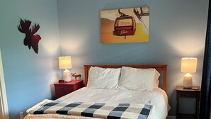 A neatly made bed with a checkered quilt, two bedside lamps, a mounted moose head, a painting of a ski lift, and a patterned rug. - Stowe 2 BR / 2 BA Vacation Condo off Mountain Road (Stowe)