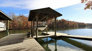 Un muelle de madera se extiende sobre un lago tranquilo, con una zona de estar cubierta y una parrilla.