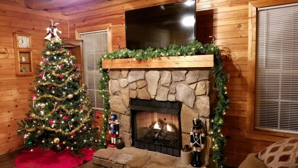Interior acogedor de una cabaña con chimenea de piedra, un árbol de Navidad y un televisor de pantalla plana montado sobre la chimenea.
