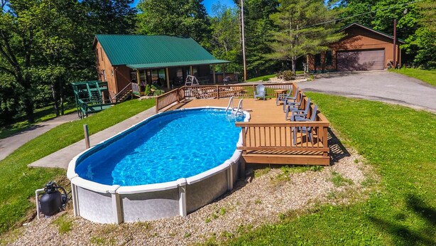 A swimming pool with a wooden deck and lounge chairs, surrounded by a grassy yard and trees. - Pines Lodge at Sand Run Lake WNF, Hocking Hills (Nelsonville)