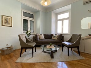 Una sala de estar con un sofá marrón, dos sillones, una mesa de centro de vidrio y una planta pequeña. Hay dos ventanas con persianas, un cuadro en la pared y una cesta en el suelo.