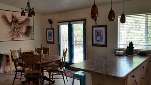 Una cocina con encimera de granito, sillas de madera, una mesa de comedor, luces colgantes y un amplio ventanal con persianas.