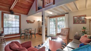 Un vaste espace de vie doté de hauts plafonds, de poutres en bois et de grandes fenêtres. On y trouve un coin repas avec une table et des chaises, ainsi qu’un coin salon avec un canapé et des fauteuils.