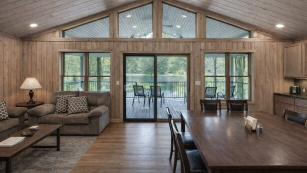 A spacious living room with a high ceiling, wooden walls, and large windows offering views of a deck and trees.