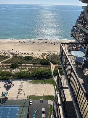 A beach view from a balcony with a tennis court and outdoor seating.
