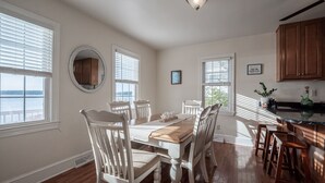 A dining area with a wooden table and white chairs, large windows with blinds, and a view of the outdoors.