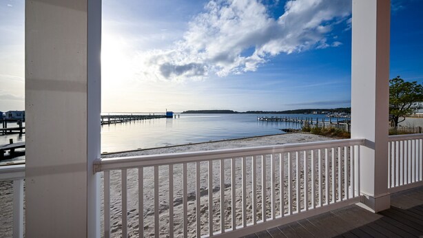 A beach view from a covered balcony with white railings, a wooden deck, and a clear sky.