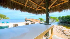 A beachside bar with a thatched roof, overlooking the sea, with lounge chairs and a wooden pier.