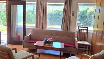 A room with a sofa, two chairs, a table, and a view of greenery through large windows.