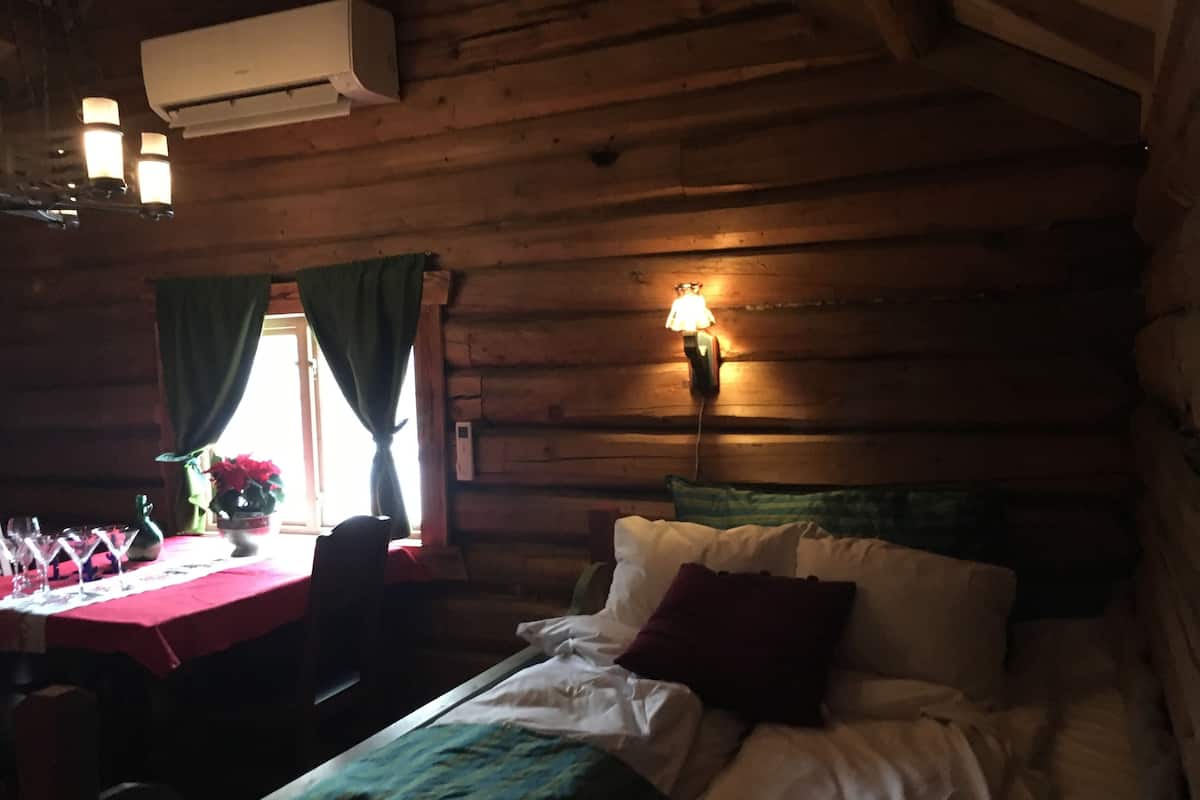 A cozy cabin-style room with a wooden interior, a bed with white and green bedding, a table set with dinnerware, and a lit wall lamp.