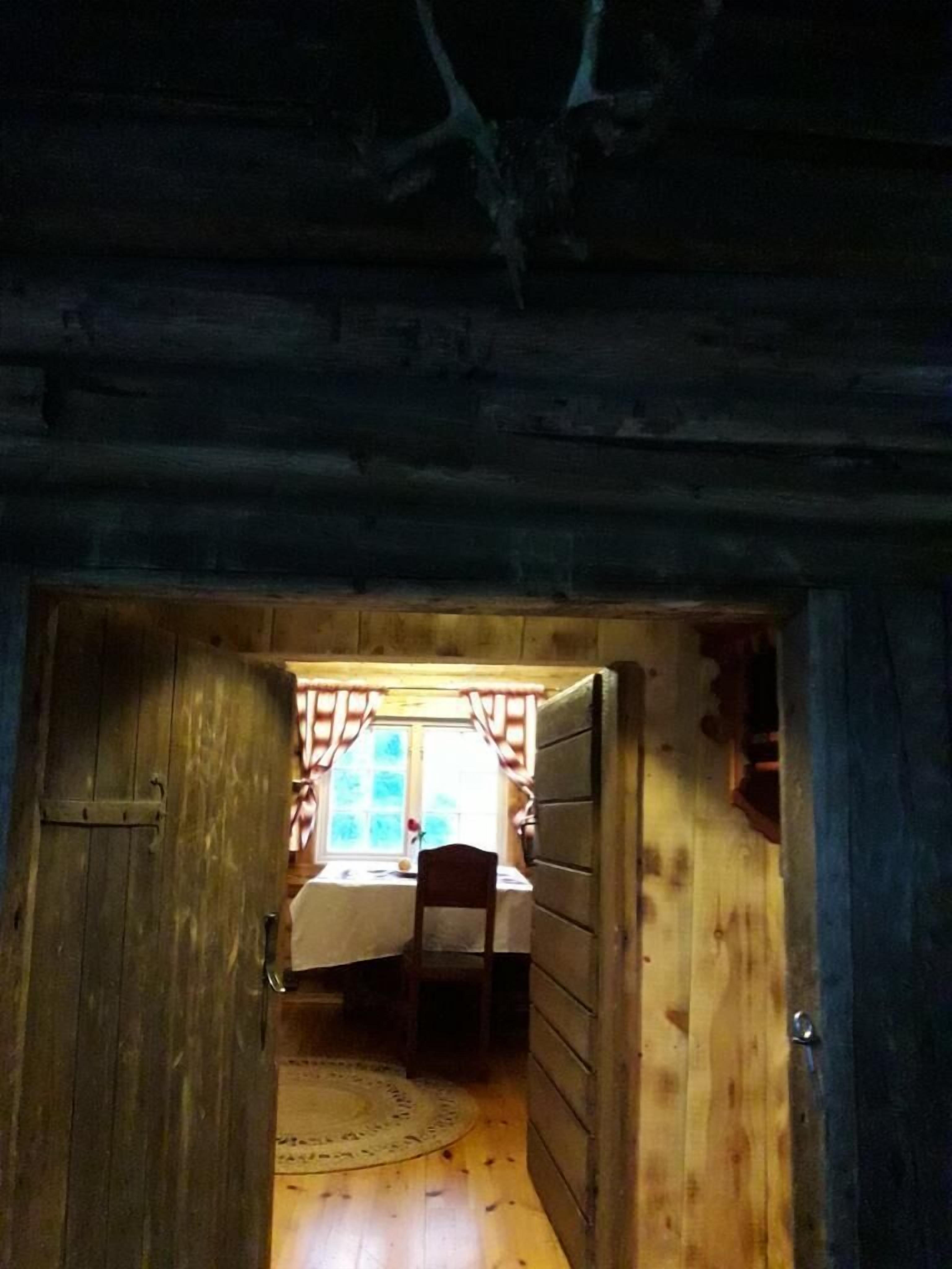 A narrow wooden hallway leading to a room with a bed, a chair, and a window with curtains.