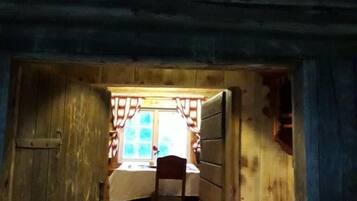 A narrow wooden hallway leading to a room with a bed, a chair, and a window with curtains.