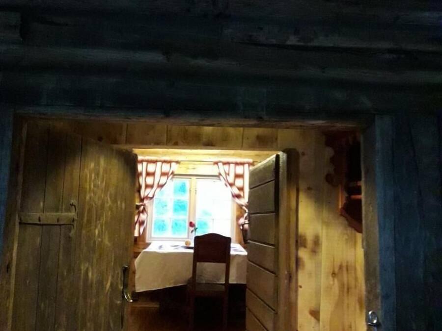 A narrow wooden hallway leading to a room with a bed, a chair, and a window with curtains.