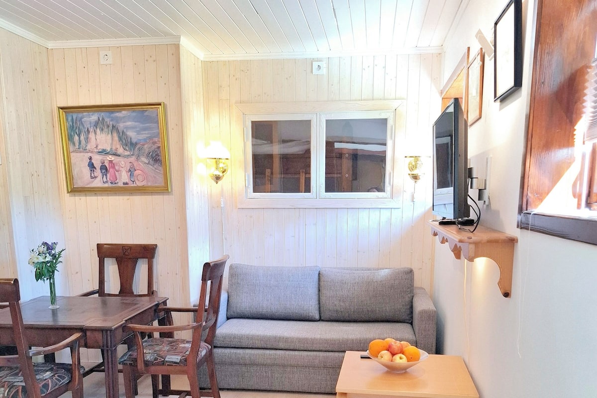 A cozy living room with a wooden floor, a grey sofa, a wooden dining table with chairs, a small wooden table with fruit, and a painting on the wall.