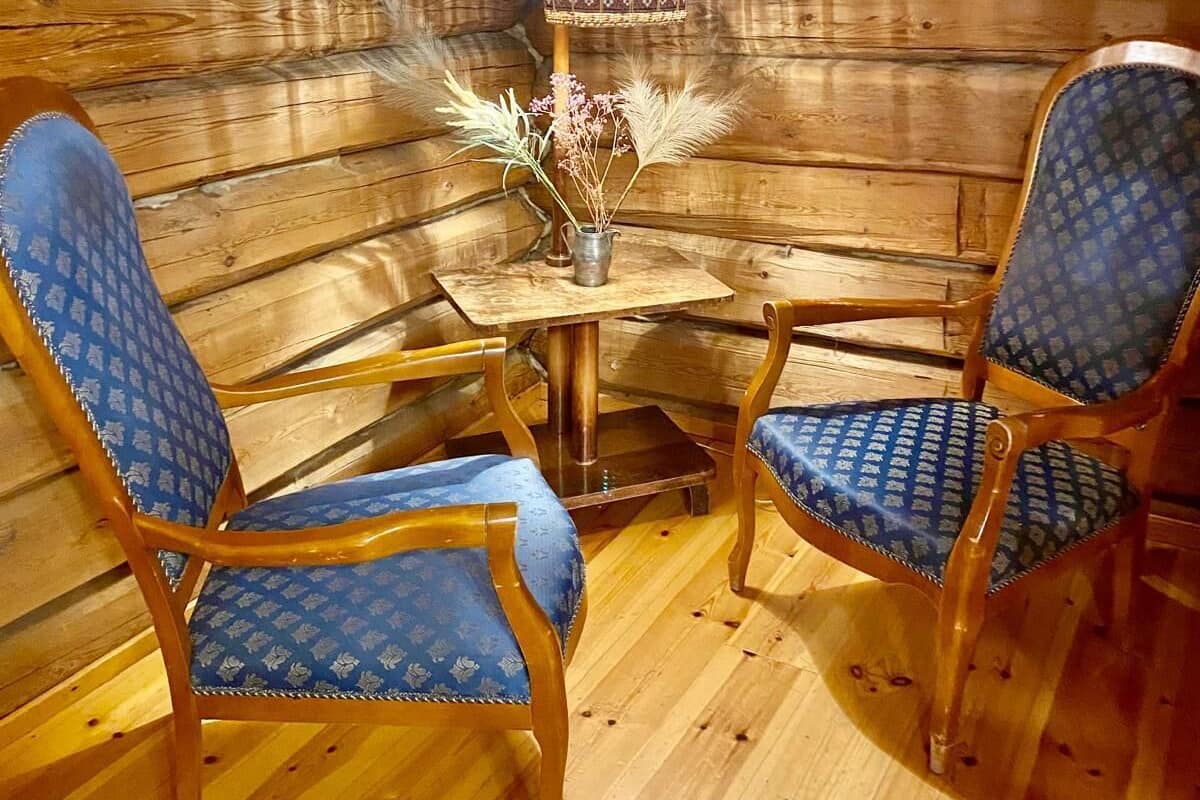 A cozy cabin interior with wooden walls, two blue-patterned chairs, a small table, and a decorative lamp.