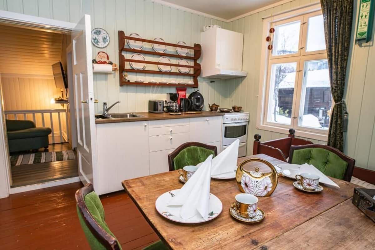 A cozy dining area with a wooden table set for tea, a kitchen with a stove and sink, and a window with curtains.
