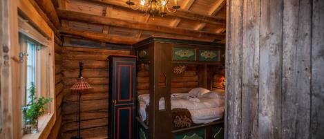 A rustic wooden cabin interior with a four-poster bed, a chandelier, and a window with plants.