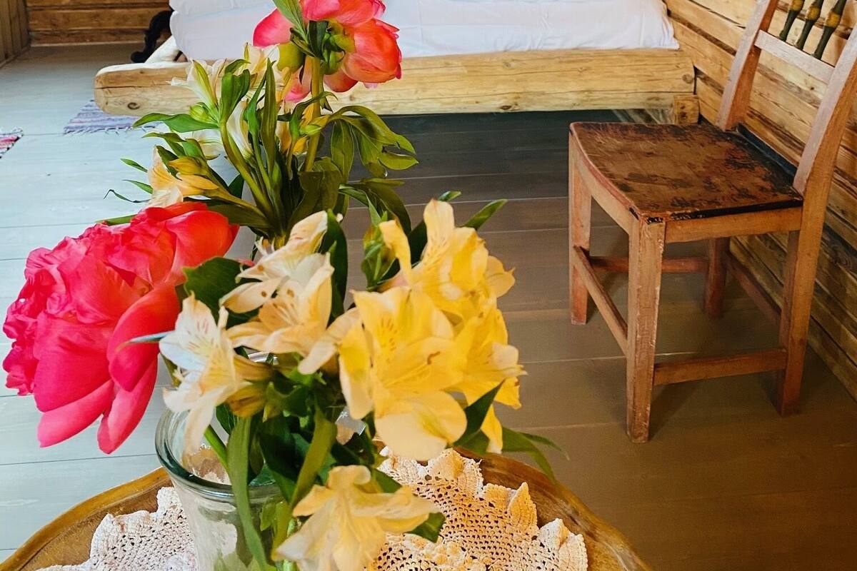 A wooden cabin room with a bed, a chair, and a table with flowers.