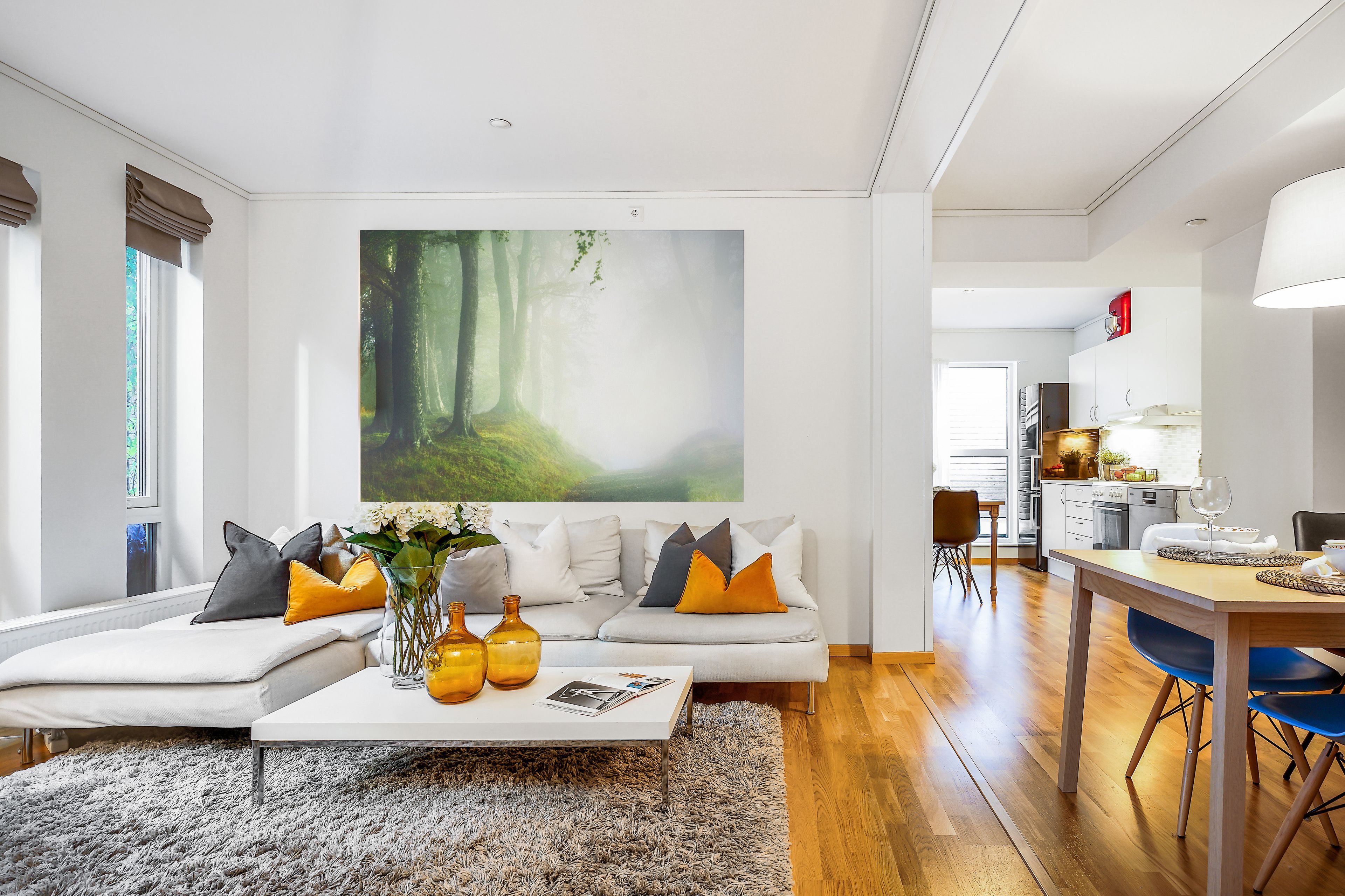 Uma sala de estar moderna com um quadro grande de uma floresta, um sofá branco com almofadas laranja e cinzentas, uma mesa de centro com vasos e uma área de jantar com mesa e cadeiras de madeira.
