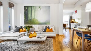 Una sala de estar moderna con un cuadro grande de un bosque, un sofá blanco con cojines naranja y grises, una mesa de centro con jarrones y un comedor con mesa y sillas de madera.