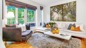Un moderno salón con un sofá gris, un sillón blanco y una mesa de centro blanca decorada con jarrones. Grandes ventanales ofrecen vistas al verde exterior.