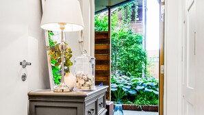 Un pasillo con un aparador de madera oscura, un espejo, una lámpara y un tarro de vidrio con monedas. Se ve una puerta que da a un jardín con plantas y una terraza de madera.