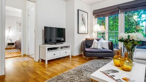 Un salón moderno con una televisión de pantalla plana sobre una consola blanca, un sofá oscuro con cojines, una mesa de centro con un jarrón y libros, y un ventanal con vistas a zonas verdes.