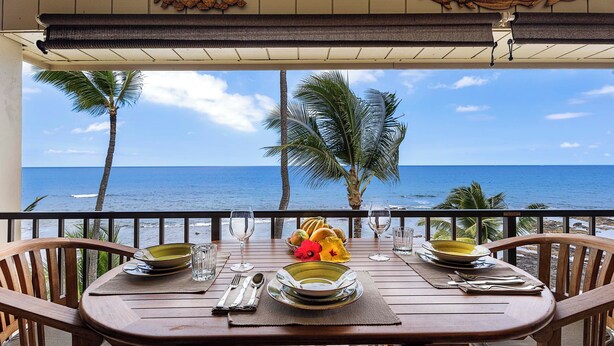 Une table en bois, dressée pour deux personnes, avec des assiettes, des verres et des couverts, donnant sur une plage bordée de palmiers et sur l’océan.