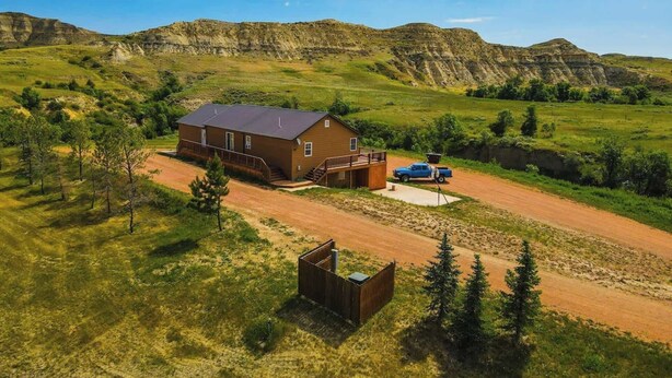 A single-story wooden cabin with a porch, a dirt driveway, and a blue pickup truck parked outside. - Boots House (Medora)