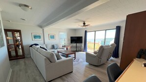 Amplia sala de estar con sofá, sillones, una mesa de centro y televisor. Hay un balcón con vista al océano.