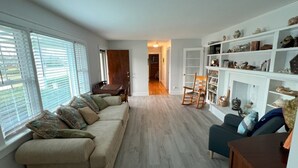 Una sala de estar con un sofá seccional color beige, un sillón oscuro y una mesa de centro de madera. Se observa una estantería blanca llena de libros y objetos decorativos.