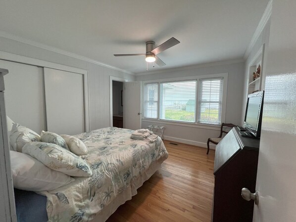 Een slaapkamer met een bed, een plafondventilator, een raam met jaloezieën en een televisie op een dressoir.