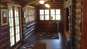 L’intérieur d’une cabane en rondins, avec une table à manger en bois, des chaises, un tapis à carreaux et une fenêtre donnant sur un paysage verdoyant.
