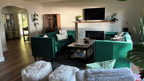 A modern living room with a fireplace, green sofas, a flat-screen TV, and a kitchen area visible through an archway. - The Bay View Brick --Stunning View of Bay 
 (Traverse City)
