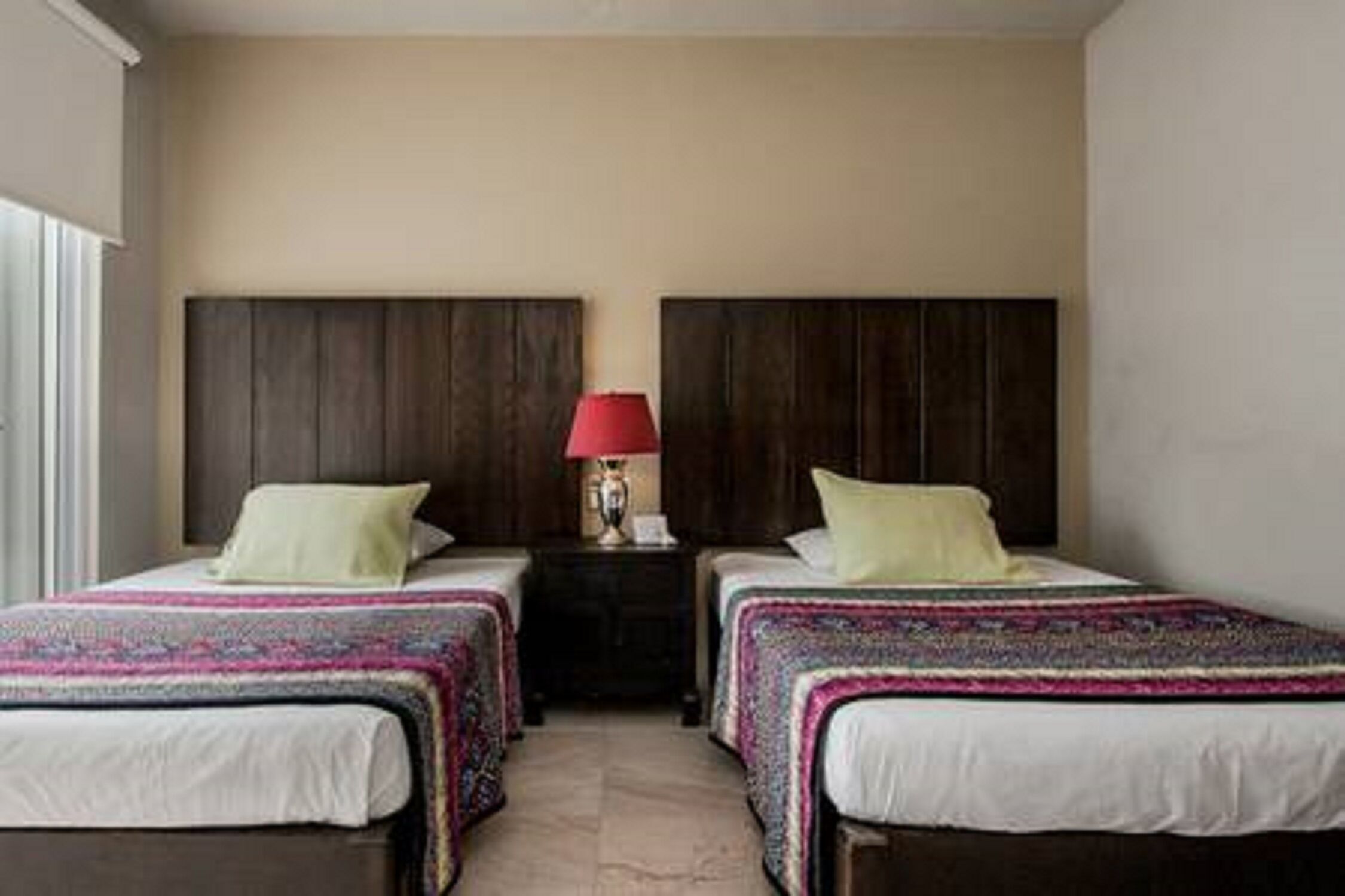 A hotel room with two single beds, a nightstand with a lamp, and a window with blinds.