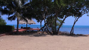 A beach with a thatched-roof structure, wooden chairs, and a sandy area. - Paradise Found!  Beautiful Private Villa On The Sea in the Village of Placencia! (Placencia Village)