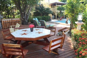 Un patio al aire libre con una mesa y sillas de madera, rodeado de abundante vegetación y una zona de piscina.