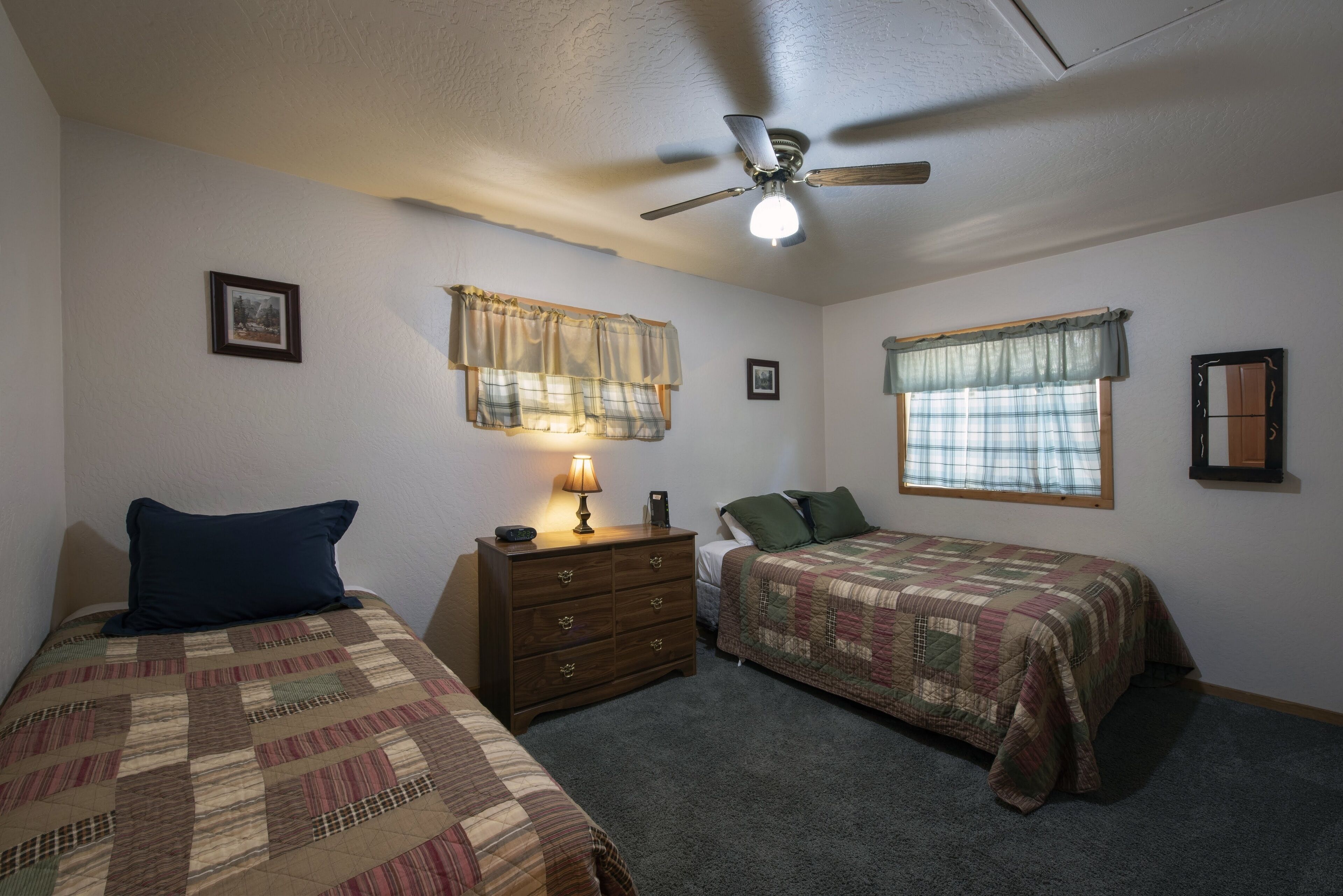 A bedroom with two beds, a dresser, a ceiling fan, and two windows with curtains.