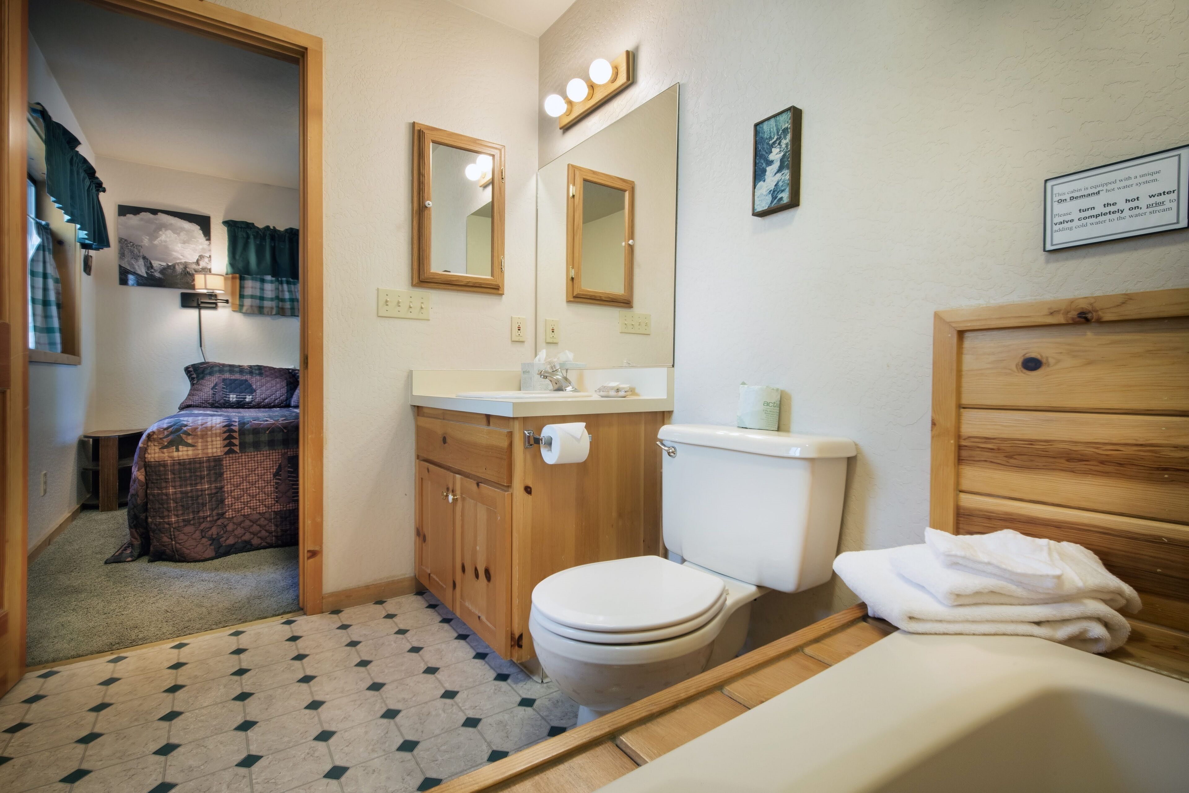Une petite salle de bain comprenant des toilettes, un lavabo et un miroir. On aperçoit un lit recouvert d’une couverture à carreaux dans la pièce adjacente.