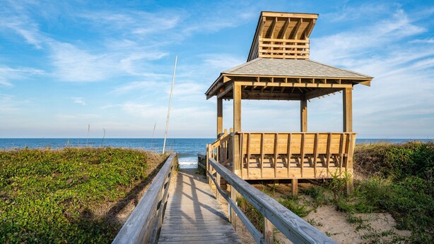 Une structure en bois installée sur la plage, dotée d’un toit incliné et d’une passerelle en bois menant à cette structure.