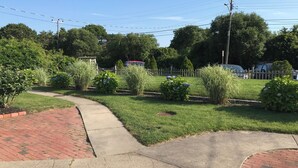 Ein gepflasterter Weg führt durch einen angelegten Garten mit verschiedenen Sträuchern und einem weißen Zaun im Hintergrund.
