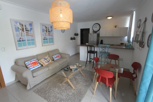 Un espace de vie compact comprenant un canapé, une table basse en verre et un coin repas avec des chaises rouges. On aperçoit une cuisine avec des meubles blancs et une horloge à l’arrière-plan.