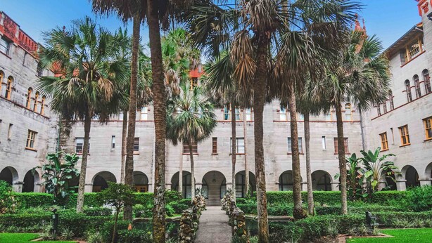 A courtyard with a central pathway flanked by palm trees and a building with arched windows and a red roof. - Ponce Landing 7 - Oceanfront Townhome - Heated Pool (Saint Augustine)