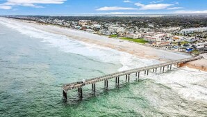An aerial view of a coastal area with a pier extending into the water, a beach, and a town in the background. - Ponce Landing 7 (Saint Augustine)