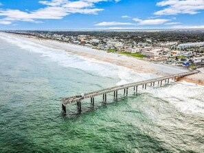 An aerial view of a coastal area with a pier extending into the water, a beach, and a town in the background. - Ponce Landing 7 (Saint Augustine)