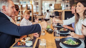 Un groupe de personnes trinquant avec des verres de vin autour d’une table de restaurant, où se trouvent de la nourriture et des boissons.